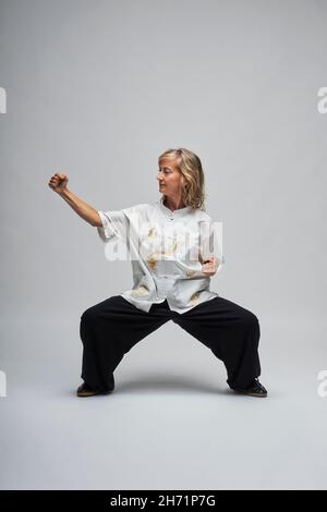 Reife blonde Frau, die Chi Kung und Tai Chi auf weißem Hintergrund praktiziert. Sie trägt eine traditionelle weiße chinesische Tai Chi Jacke, schwarze Hosen und BL Stockfoto