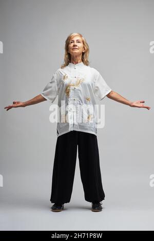 Reife blonde Frau, die Chi Kung und Tai Chi auf weißem Hintergrund praktiziert. Sie trägt eine traditionelle weiße chinesische Tai Chi Jacke, schwarze Hosen und BL Stockfoto
