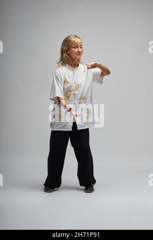 Reife blonde Frau, die Chi Kung und Tai Chi auf weißem Hintergrund praktiziert. Sie trägt eine traditionelle weiße chinesische Tai Chi Jacke, schwarze Hosen und BL Stockfoto