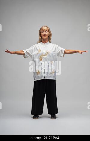 Reife blonde Frau, die Chi Kung und Tai Chi auf weißem Hintergrund praktiziert. Sie trägt eine traditionelle weiße chinesische Tai Chi Jacke, schwarze Hosen und BL Stockfoto