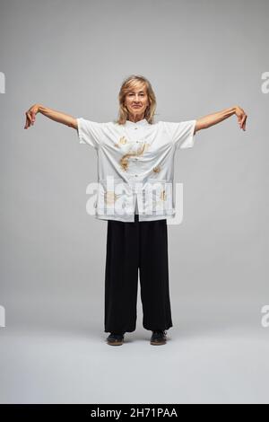 Reife blonde Frau, die Chi Kung und Tai Chi auf weißem Hintergrund praktiziert. Sie trägt eine traditionelle weiße chinesische Tai Chi Jacke, schwarze Hosen und BL Stockfoto