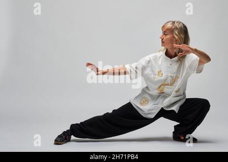 Reife blonde Frau, die Chi Kung und Tai Chi auf weißem Hintergrund praktiziert. Sie trägt eine traditionelle weiße chinesische Tai Chi Jacke, schwarze Hosen und BL Stockfoto