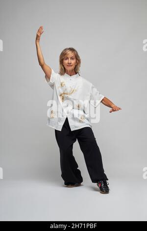 Reife blonde Frau, die Chi Kung und Tai Chi auf weißem Hintergrund praktiziert. Sie trägt eine traditionelle weiße chinesische Tai Chi Jacke, schwarze Hosen und BL Stockfoto