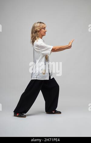 Reife blonde Frau, die Chi Kung und Tai Chi auf weißem Hintergrund praktiziert. Sie trägt eine traditionelle weiße chinesische Tai Chi Jacke, schwarze Hosen und BL Stockfoto