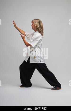 Reife blonde Frau, die Chi Kung und Tai Chi auf weißem Hintergrund praktiziert. Sie trägt eine traditionelle weiße chinesische Tai Chi Jacke, schwarze Hosen und BL Stockfoto