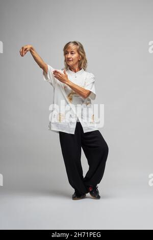 Reife blonde Frau, die Chi Kung und Tai Chi auf weißem Hintergrund praktiziert. Sie trägt eine traditionelle weiße chinesische Tai Chi Jacke, schwarze Hosen und BL Stockfoto