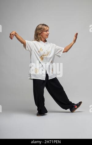 Reife blonde Frau, die Chi Kung und Tai Chi auf weißem Hintergrund praktiziert. Sie trägt eine traditionelle weiße chinesische Tai Chi Jacke, schwarze Hosen und BL Stockfoto
