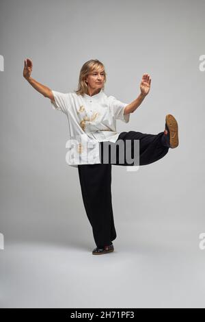 Reife blonde Frau, die Chi Kung und Tai Chi auf weißem Hintergrund praktiziert. Sie trägt eine traditionelle weiße chinesische Tai Chi Jacke, schwarze Hosen und BL Stockfoto