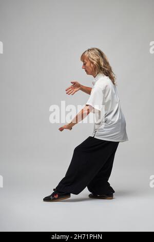 Reife blonde Frau, die Chi Kung und Tai Chi auf weißem Hintergrund praktiziert. Sie trägt eine traditionelle weiße chinesische Tai Chi Jacke, schwarze Hosen und BL Stockfoto