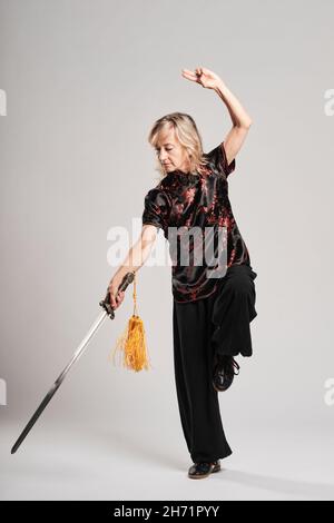 Reife blonde Frau, die Tai Chin mit Schwert auf weißem Hintergrund praktiziert, trägt eine traditionelle chinesische schwarze Jacke mit roter chinesischer Dekoration, schwarz Stockfoto
