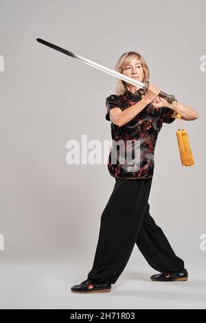 Reife blonde Frau, die Tai Chin mit Schwert auf weißem Hintergrund praktiziert, trägt eine traditionelle chinesische schwarze Jacke mit roter chinesischer Dekoration, schwarz Stockfoto