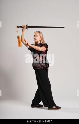 Reife blonde Frau, die Tai Chin mit Schwert auf weißem Hintergrund praktiziert, trägt eine traditionelle chinesische schwarze Jacke mit roter chinesischer Dekoration, schwarz Stockfoto