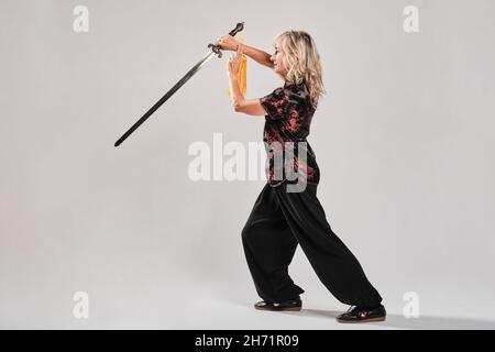 Reife blonde Frau, die Tai Chin mit Schwert auf weißem Hintergrund praktiziert, trägt eine traditionelle chinesische schwarze Jacke mit roter chinesischer Dekoration, schwarz Stockfoto