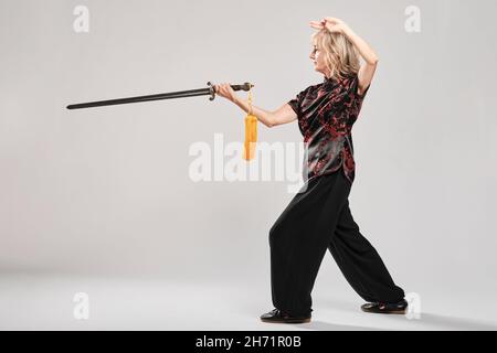Reife blonde Frau, die Tai Chin mit Schwert auf weißem Hintergrund praktiziert, trägt eine traditionelle chinesische schwarze Jacke mit roter chinesischer Dekoration, schwarz Stockfoto