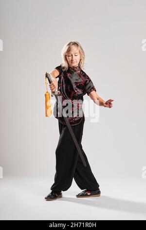 Reife blonde Frau, die Tai Chin mit Schwert auf weißem Hintergrund praktiziert, trägt eine traditionelle chinesische schwarze Jacke mit roter chinesischer Dekoration, schwarz Stockfoto