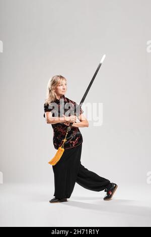 Reife blonde Frau, die Tai Chin mit Schwert auf weißem Hintergrund praktiziert, trägt eine traditionelle chinesische schwarze Jacke mit roter chinesischer Dekoration, schwarz Stockfoto