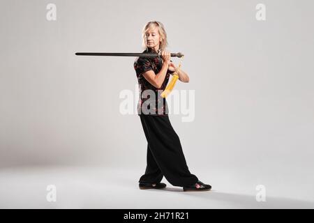 Reife blonde Frau, die Tai Chin mit Schwert auf weißem Hintergrund praktiziert, trägt eine traditionelle chinesische schwarze Jacke mit roter chinesischer Dekoration, schwarz Stockfoto