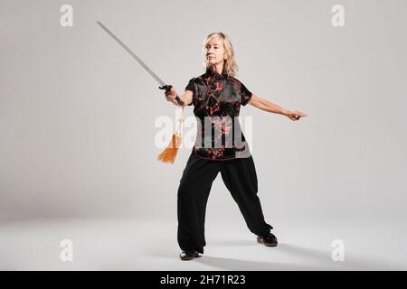 Reife blonde Frau, die Tai Chin mit Schwert auf weißem Hintergrund praktiziert, trägt eine traditionelle chinesische schwarze Jacke mit roter chinesischer Dekoration, schwarz Stockfoto