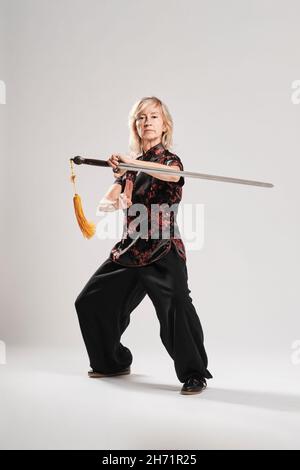 Reife blonde Frau, die Tai Chin mit Schwert auf weißem Hintergrund praktiziert, trägt eine traditionelle chinesische schwarze Jacke mit roter chinesischer Dekoration, schwarz Stockfoto