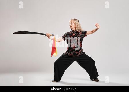 Reife blonde Frau, die Tai Chin mit Säbel auf weißem Hintergrund praktiziert, trägt eine traditionelle chinesische schwarze Jacke mit roter chinesischer Dekoration, schwarz Stockfoto