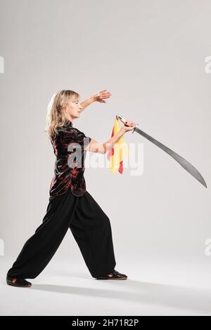 Reife blonde Frau, die Tai Chin mit Säbel auf weißem Hintergrund praktiziert, trägt eine traditionelle chinesische schwarze Jacke mit roter chinesischer Dekoration, schwarz Stockfoto