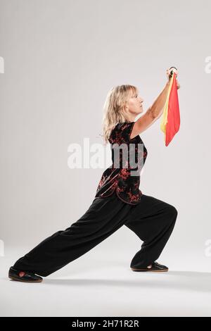 Reife blonde Frau, die Tai Chin mit Säbel auf weißem Hintergrund praktiziert, trägt eine traditionelle chinesische schwarze Jacke mit roter chinesischer Dekoration, schwarz Stockfoto