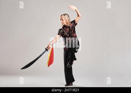 Reife blonde Frau, die Tai Chin mit Säbel auf weißem Hintergrund praktiziert, trägt eine traditionelle chinesische schwarze Jacke mit roter chinesischer Dekoration, schwarz Stockfoto