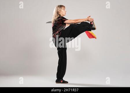 Reife blonde Frau, die Tai Chin mit Säbel auf weißem Hintergrund praktiziert, trägt eine traditionelle chinesische schwarze Jacke mit roter chinesischer Dekoration, schwarz Stockfoto