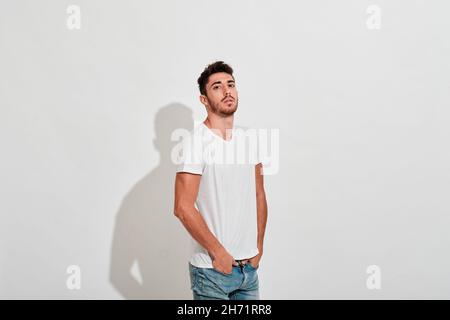 Junger Mann, der Jeans und einen weißen Scheiß vor einer weißen Wand trägt, während er auf die Kamera schaut. Stockfoto
