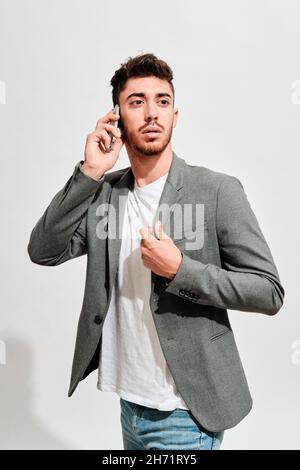 Ernsthafter junger Mann in Jeans, einem weißen T-Shirt und einem grauen Blazer, der vor einer weißen Wand läuft, während er mit jemandem auf seinem Mobiltelefon spricht Stockfoto