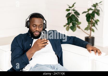 Attraktiver, tausendjähriger junger bärtiger Afrikaner, der Kopfhörer trägt, sich entspannt und auf dem bequemen Sofa liegend, Musik hört, mit Freunden in sozialen Medien auf dem Mobiltelefon chattet und online datet Stockfoto