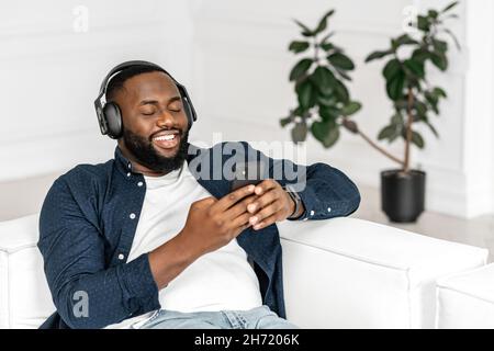 Zufriedener, gutaussehender afrikanischer Mann, der Smartphone und kabellose Kopfhörer zum Musikhören verwendet, legt sich schwarzer Mann auf die Couch, die Augen sind in Träumen geschlossen, genießt Lieblingstitel und ruht sich in gemütlicher Atmosphäre aus Stockfoto