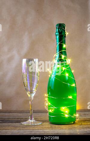 Eine Flasche grünen Champagner, umhüllt von einer glühenden Girlande wie ein Weihnachtsbaum und Glas. Neues Jahr Konzept auf natürlichem Hintergrund Stockfoto