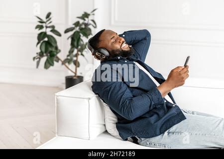 Junger afroamerikanischer Mann mit lässigem Hemd sitzt auf weißem Sofa in moderner Wohnung, hört Musik auf dem Smartphone, hört Hörbücher, Podcast, schwarzer Mann, der zu Hause ruht, mit geschlossenen Augen liegt Stockfoto