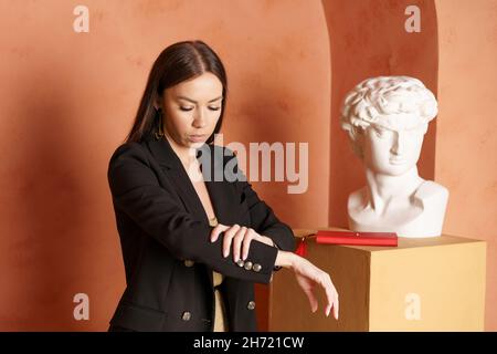 Porträt von glücklich lächelnden Brünette Geschäftsfrau in schwarzer Jacke und goldenes Kleid zuversichtlich posiert mit Gips Büste Figur im Studio auf dem Hintergrund. Business-Studio-Konzept. Stockfoto