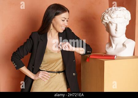 Die Geschäftsfrau steht im Studio neben dem Gipskopf. Kaukasische Mädchen und Gips Büste. Lehrer oder Kunstkritiker in Kleid und schwarzer Jacke Stockfoto