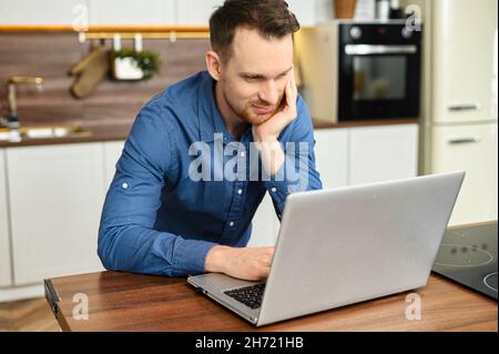 Fokussierter freiberuflicher Mann in elegantem Casual-Shirt mit Laptop in der Küche zu Hause. Lächelnder Mann, der aus der Ferne arbeitet und online Nachrichten veröffentlicht Stockfoto