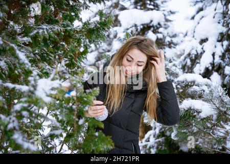 Glückliches Mädchen in warmen Winterkleidung im verschneiten Wald hängt Weihnachtsbälle auf weihnachtsbaum. Kaukasische Frau schmückt weihnachtsbaum im Winter verschneiten Park in warmer Kleidung Stockfoto