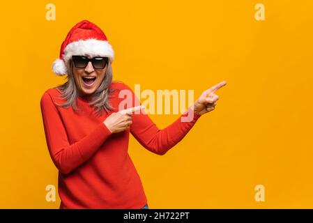 Staunende grauhaarige Frau in weihnachtsmann Hut und Sonnenbrille stehend isoliert über gelbem Hintergrund zeigt mit den Fingern auf den leeren Raum für Werbung, Mockup Bild, überprüfen Sie dies, lustiges Konzept Stockfoto