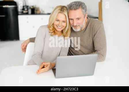 Der moderne, fröhliche, reife Mann und seine Frau sitzen am Schreibtisch in der Küche, mit einem Laptop am Bildschirm und einem lächelnden, glücklichen Paar mittleren Alters, das Filme guckt, im Internet surft und Videoanruf abgibt Stockfoto