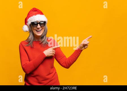 Freundliche und lächelnde ältere Frau mit Weihnachtsmann-Hut und Sonnenbrille zeigt die Finger zur Seite auf leeren Raum, Platz für Werbetexte kopieren, reife grauhaarige Frau mit rotem Pullover isoliert auf Gelb Stockfoto