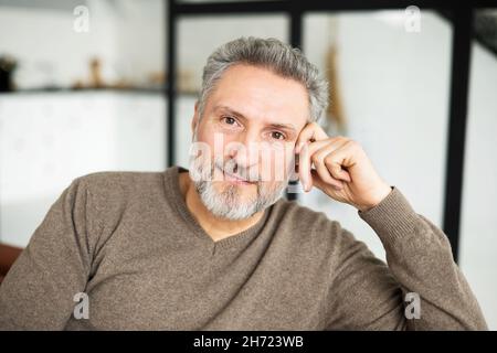 Porträt eines attraktiven, charismatischen, reifen Mannes, der die Kamera anschaut. Ein hübscher grauhaariger bärtiger Rüde in legerer Kleidung sitzt in entspannter Pose im Innenbereich, mit Kopfschuss Stockfoto