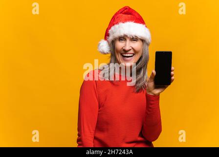 Begeistert von dem Urlaubsangebot reife Frau in santa hat hält Smartphone mit leerem Bildschirm, überglücklich mittleren Alters Dame demonstriert Telefon mit leerem Display, isoliert auf gelb Stockfoto
