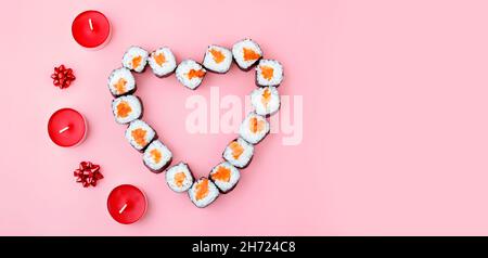 Lachs-Sushi-Rollen in Herzform, neben roten Kerzen und einem rosa Hintergrund. Das Konzept der japanischen Küche zum Valentinstag Stockfoto