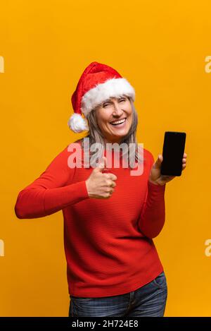 Vertikales Porträt einer urkomischen älteren Frau im roten weihnachtsmann-Hut mit Smartphone und leerem Bildschirm, isoliert auf gelber, grauhaariger Dame mittleren Alters, die den Daumen nach oben zeigt, wie ein Schild Stockfoto