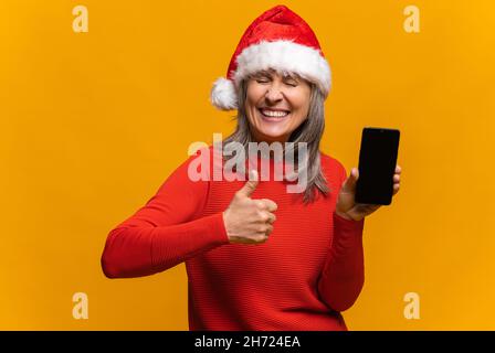 Porträt einer urkomischen älteren Frau mit faltenem Gesicht und geschlossenen Augen im weihnachtsmann-Hut, das Smartphone mit leerem Bildschirm hält, isoliert auf einer gelben, grauhaarigen Dame mittleren Alters, die den Daumen nach oben zeigt, wie ein Schild Stockfoto