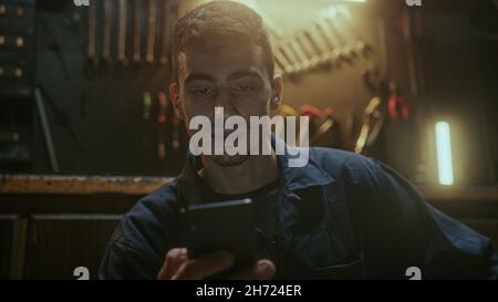 Junger, hübscher Schmied, der vor der Werkbank sitzt, sich ausruht und Smartphone benutzt Stockfoto
