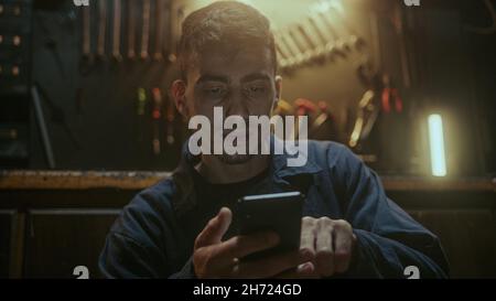 Junger, hübscher Schmied, der vor der Werkbank sitzt, sich ausruht und Smartphone benutzt Stockfoto