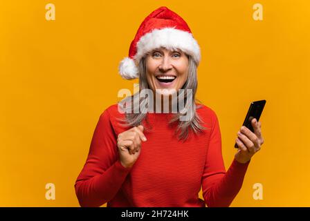 Im mittleren Alter glücklich schockiert Frau in santa Hut hält Smartphone und Blick auf die Kamera mit überrascht geöffneten Mund, verwirrt fröhlich reifen Weibchen, isoliert auf gelb. Werbekonzept für das neue Jahr Stockfoto