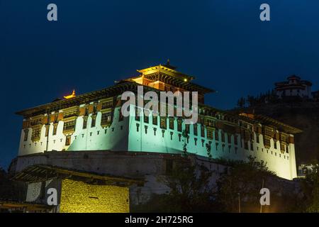 Die Rinpung oder Rinchen Pung Dzong beleuchtet in der Nacht in Paro, Bhutan. Der Wachturm Ta Dzong befindet sich oben rechts. Stockfoto